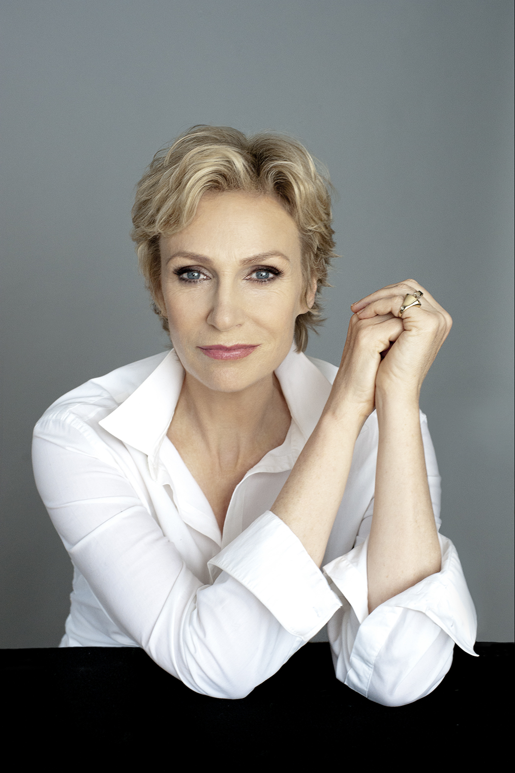 Jane Lynch wears a collared white shirt and poses with her arms folded next to her face